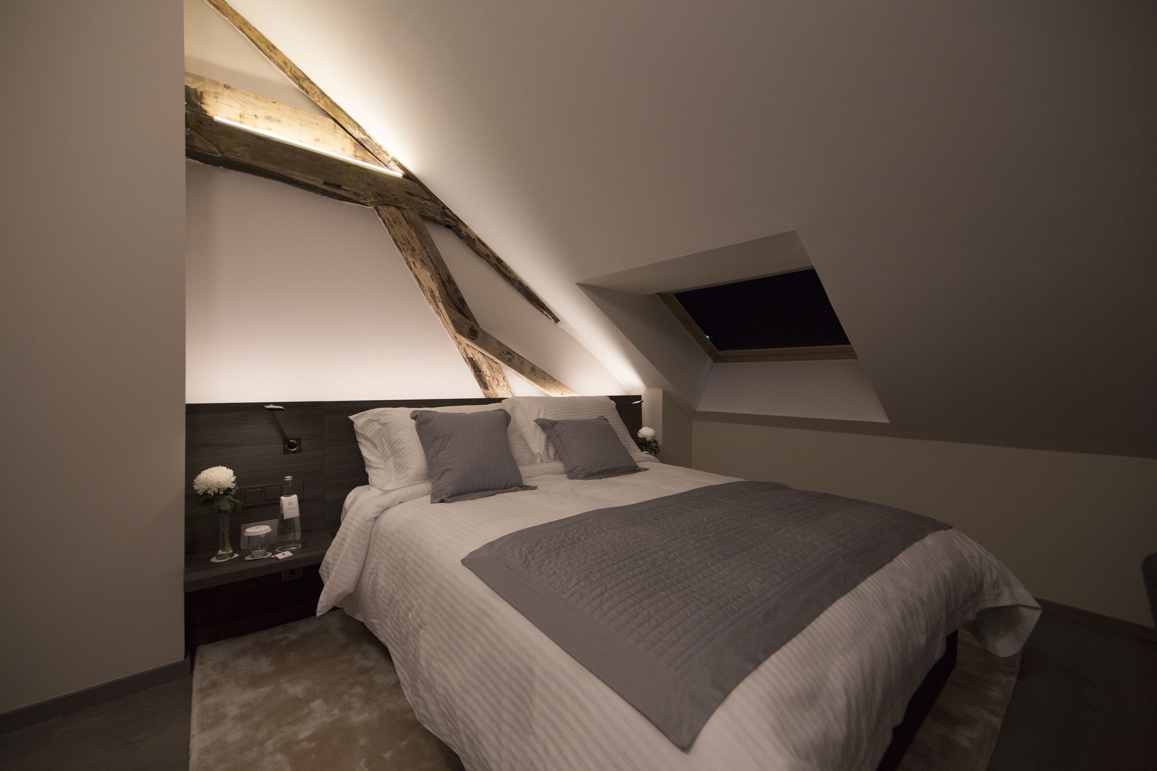 Een slaapkamer met een schuine kap, dakraam, bed met grijze beddengoed en nachtkastje met vaas en fles.