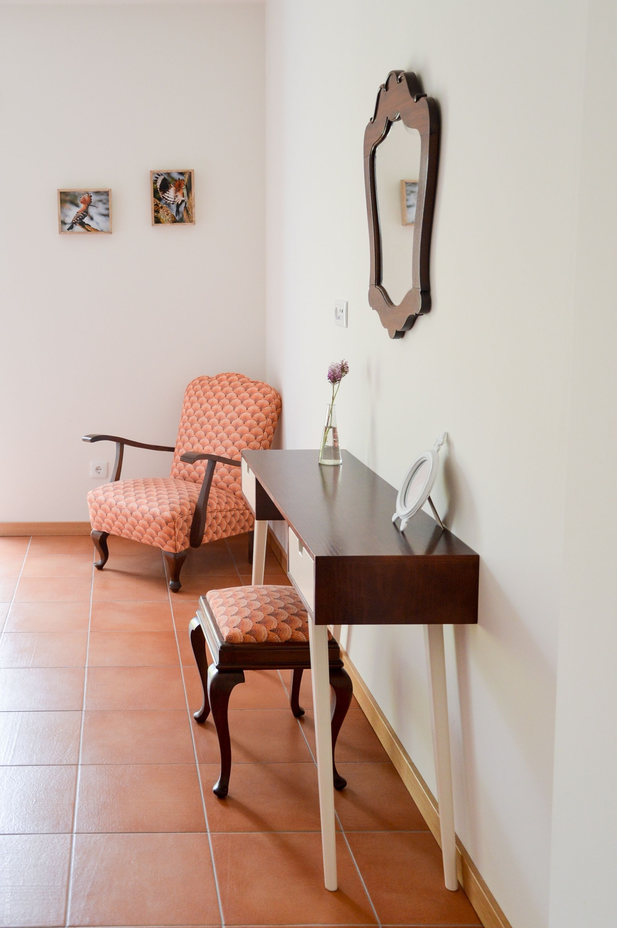 A room with a wooden chair, a small table, a mirror, and two framed pictures on the wall.