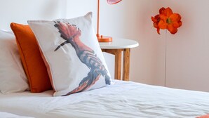A neatly made bed with white and orange bedding, a small round table with a lamp, and a single red flower on the wall. - Retiro do Bosque Country House (Alcanena)