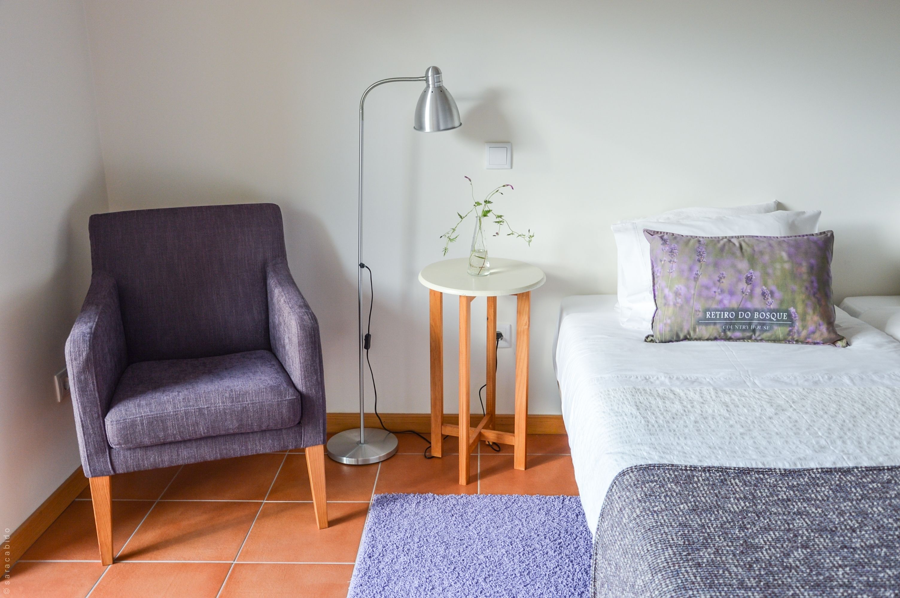 A room with a grey armchair, a small round table with a plant, a floor lamp, and a bed with a white pillow and a purple floral pillow.