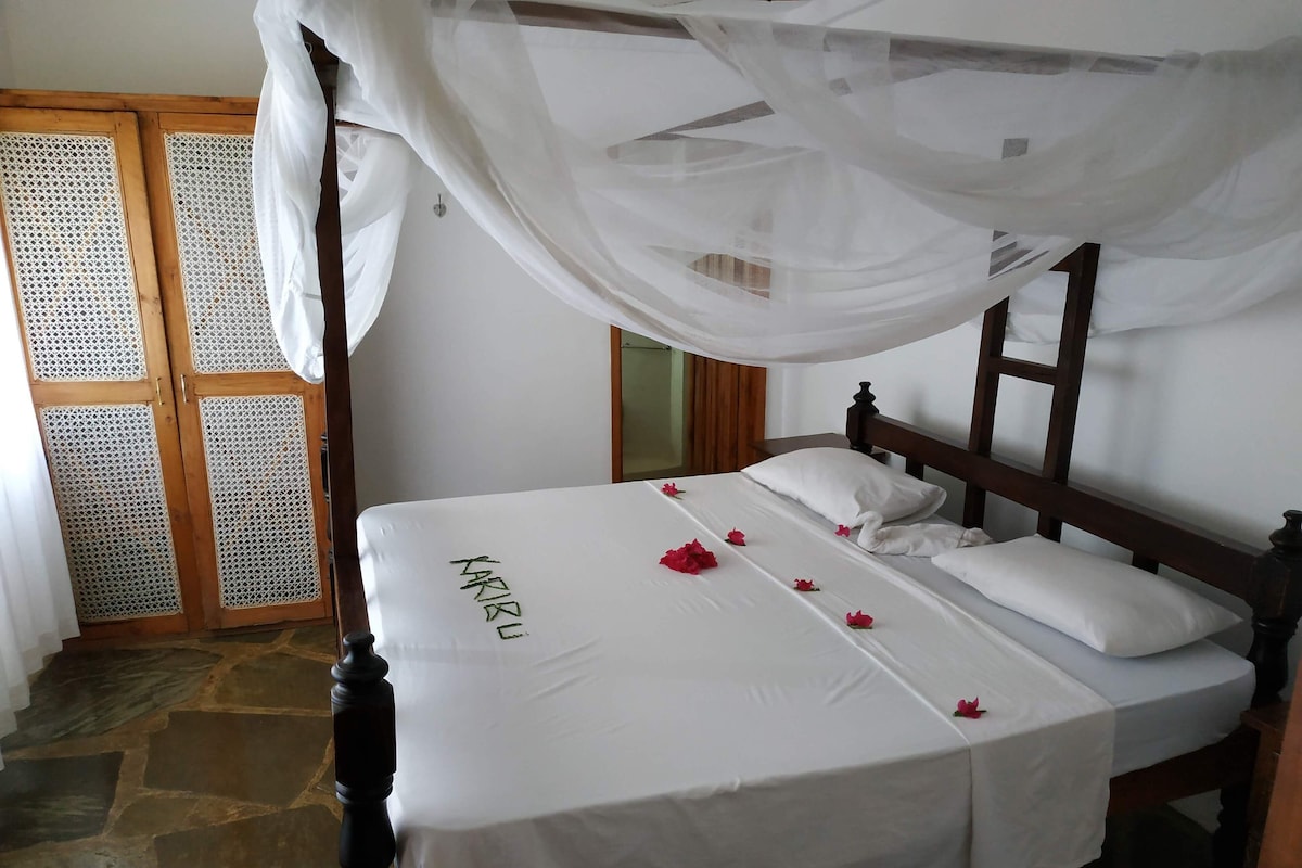 Una cama con dosel, suelo de piedra y puerta de madera con mosquitera.
