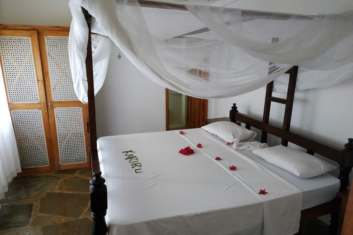 Una cama con dosel, suelo de piedra y puerta de madera con mosquitera.