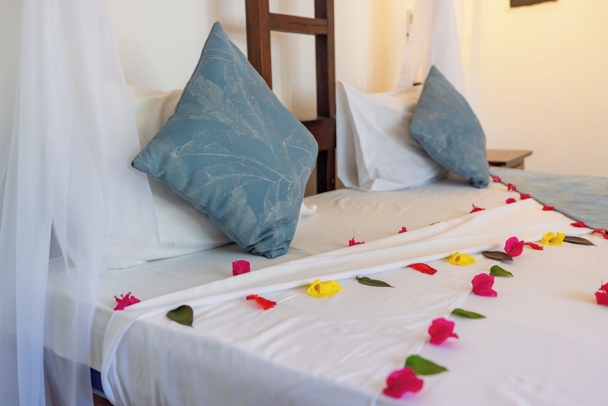 Una cama con ropa de cama blanca, almohadas azules y pétalos de flores esparcidos.