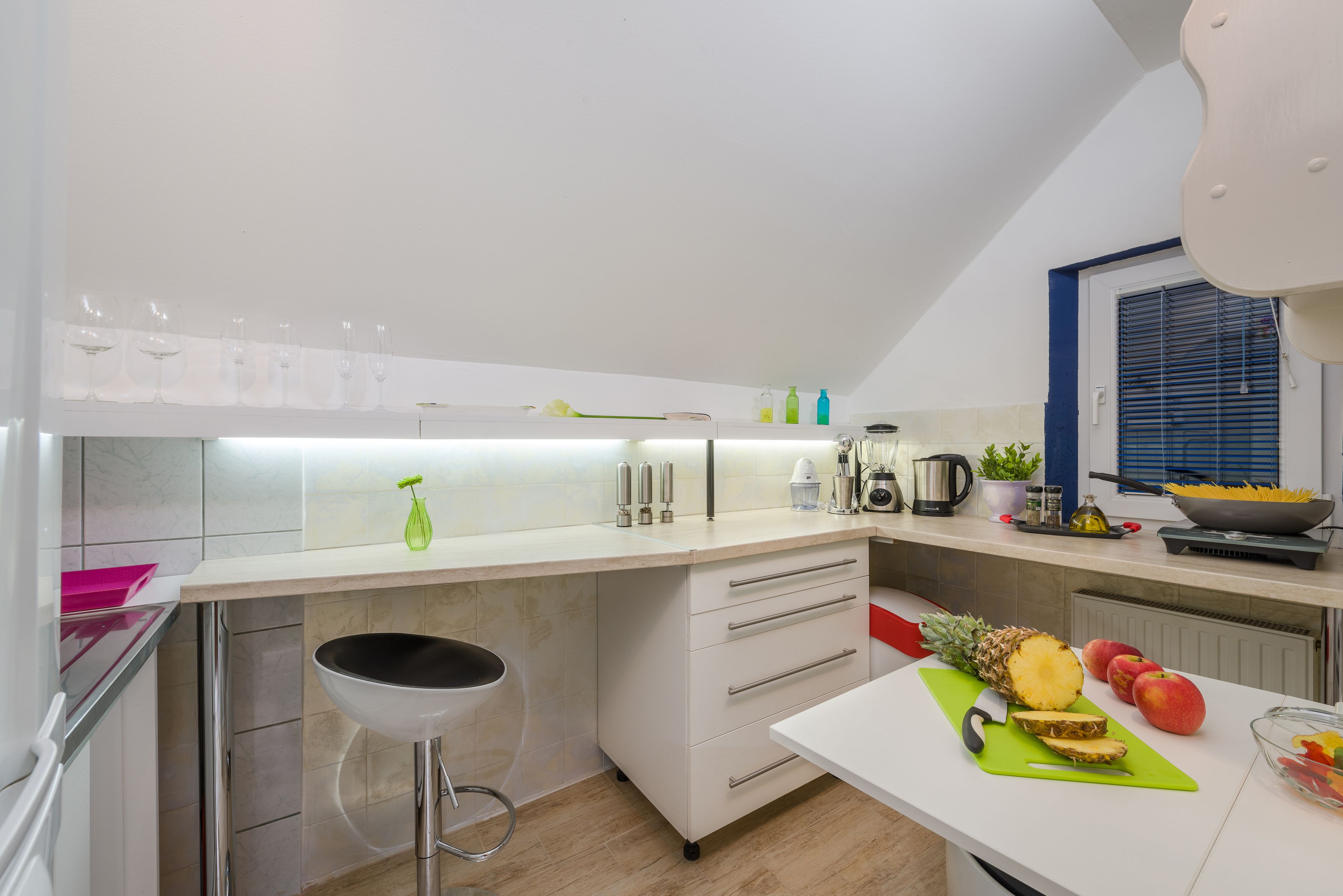 Una cocina moderna con encimera blanca, un taburete, una tabla de cortar con piña y manzanas, y un cuenco de fruta.