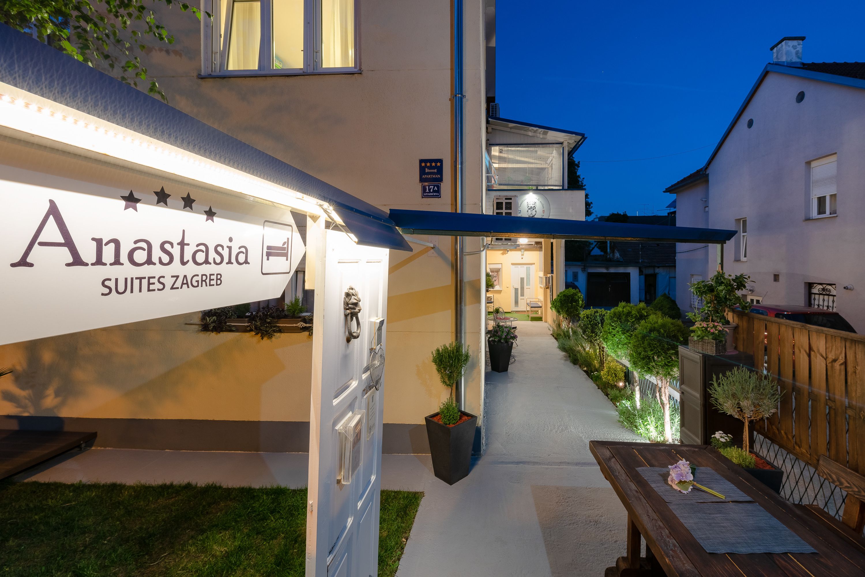 A hotel entrance with a sign for Anastasia Suites Zagreb, a pathway leading to the entrance, and outdoor seating area with a table and chairs.