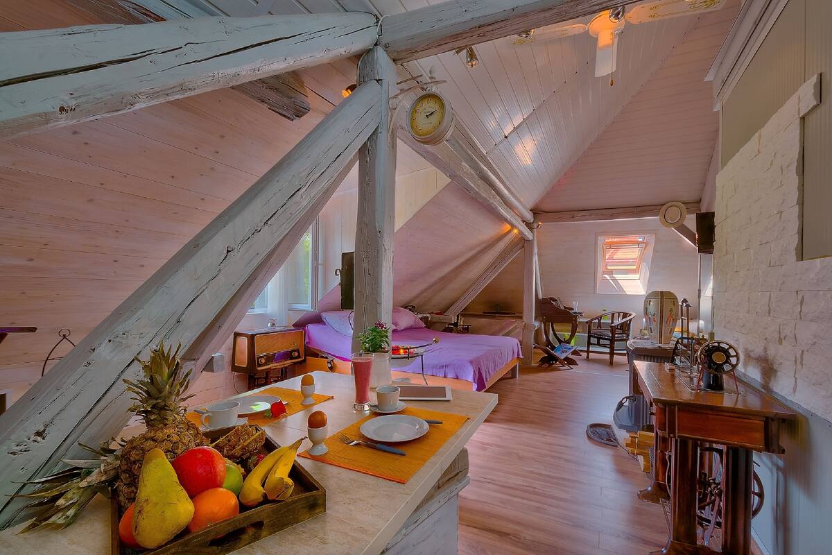 A rustic attic bedroom with a sloped ceiling, a bed, a dining table with breakfast items, and a vintage sewing machine.