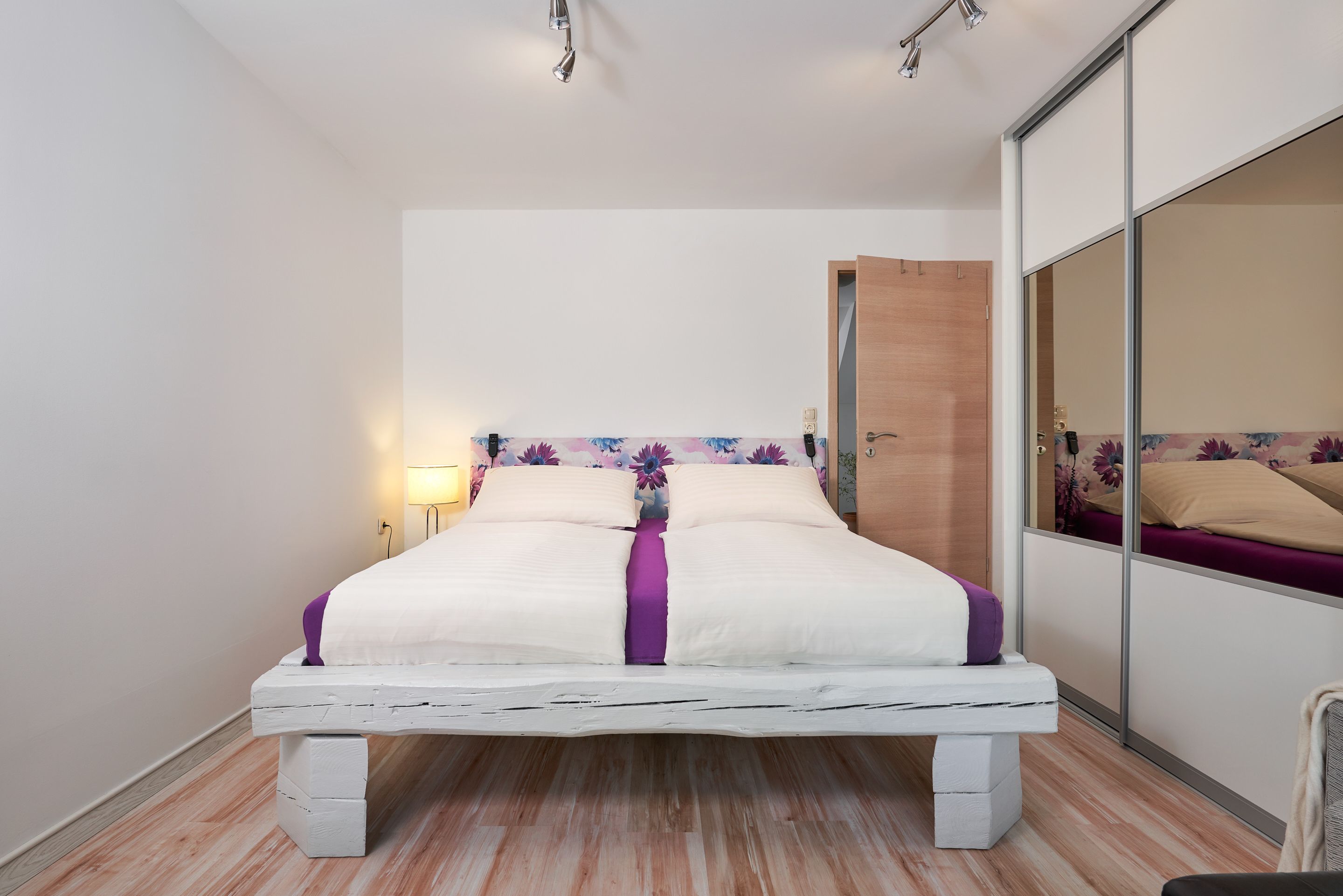A modern bedroom with a white bed, purple accents, a wooden headboard, and a large mirror.