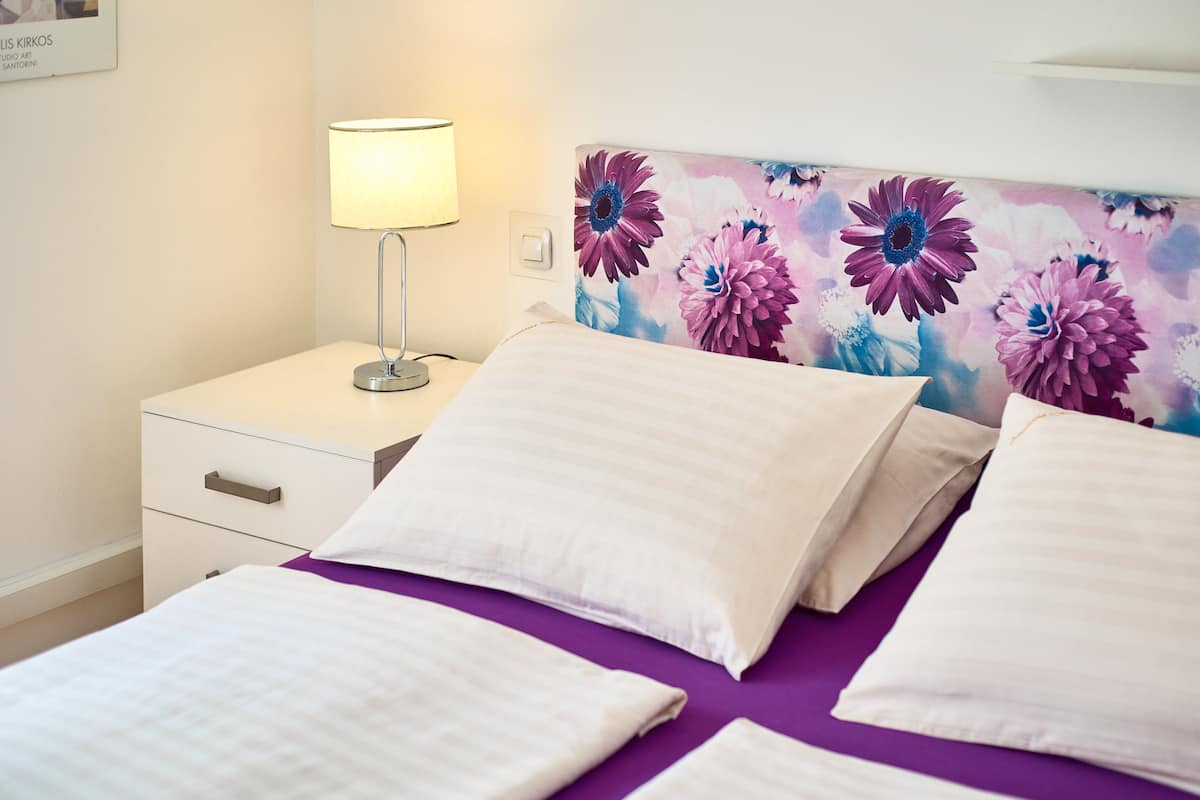 A neatly made bed with white linens, a bedside table with a lamp, and a floral headboard.