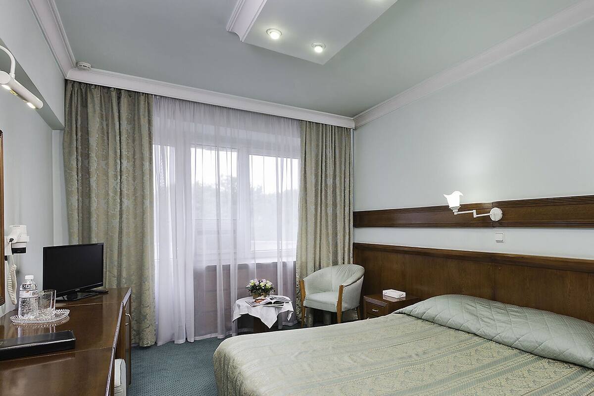A hotel room with a bed, a desk with a computer, a chair, a small table with a vase, and a window with curtains.