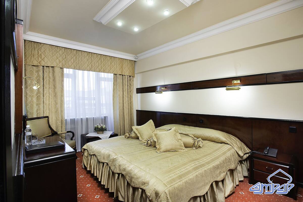 A hotel room with a large bed, a bedside table, a nightstand, a mirror, and a window with curtains.