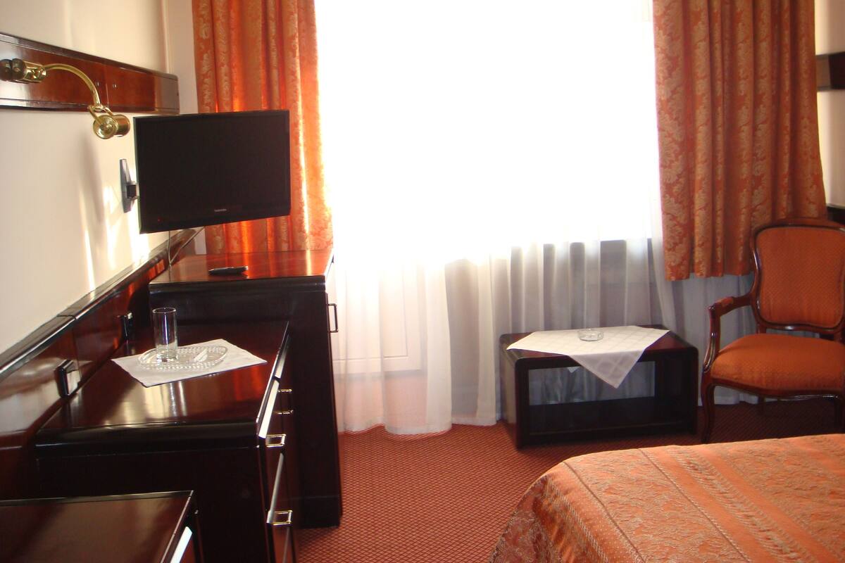 A hotel room with a bed, a desk with a television, a chair, and a window with curtains.