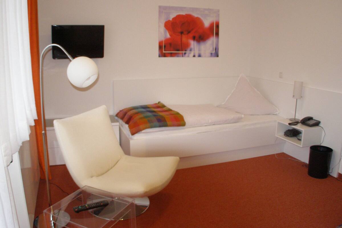 A small, modern room with a white bed, a white chair, a small table with a remote control, a lamp, and a painting on the wall.