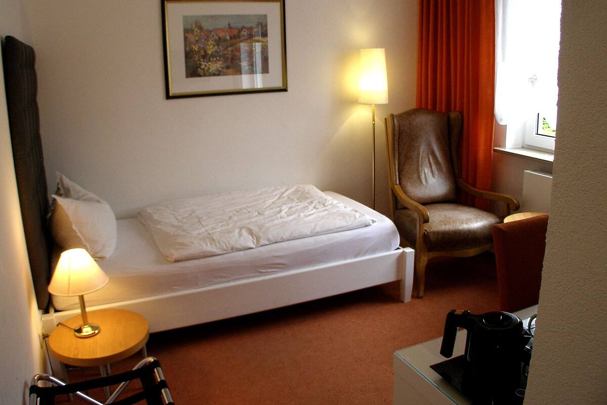A hotel room with a single bed, a chair, a lamp, and a framed picture on the wall.