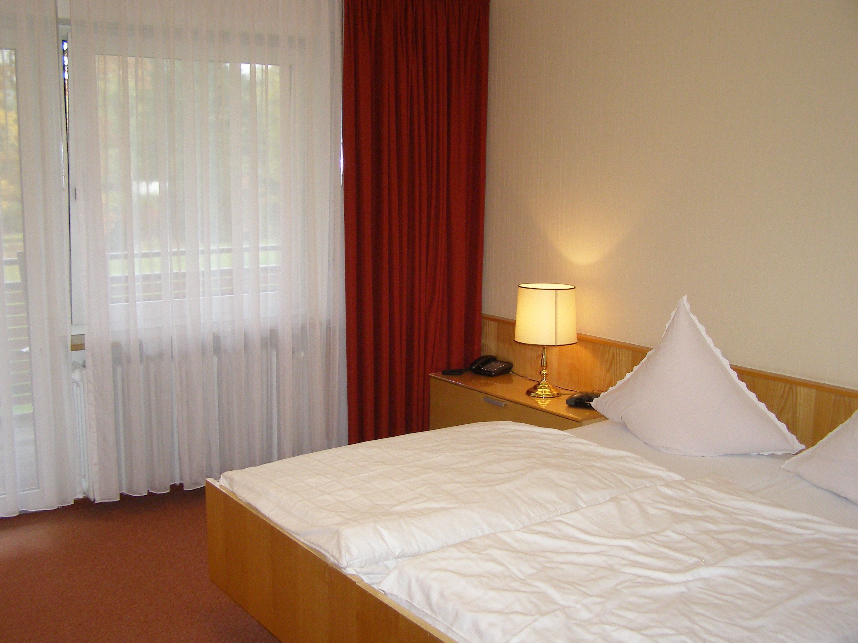 A hotel room with a bed, a nightstand with a lamp, a telephone, and a window with curtains.