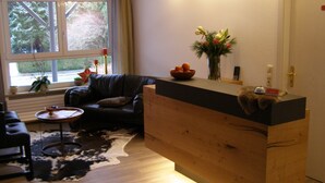 A living room with a black leather sofa, a wooden coffee table, and a cowhide rug. There is a window with a view of trees and a vase with flowers on a shelf. - Haus Katharina - Hotel am Park (Bad Steben)