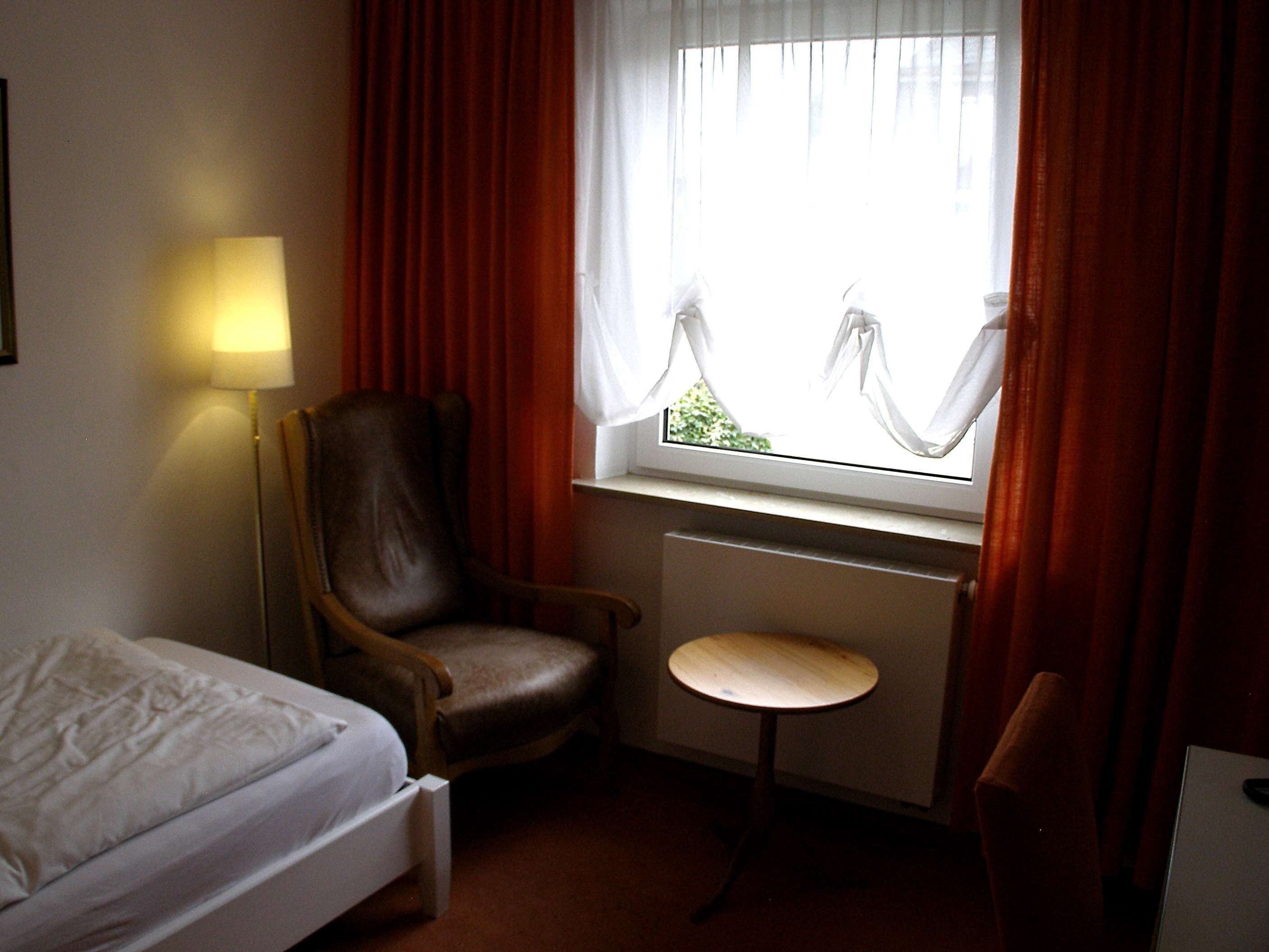 Ein Hotelzimmer mit Bett, Sessel, kleinem Tisch, Lampe und einem Fenster mit Vorhängen.