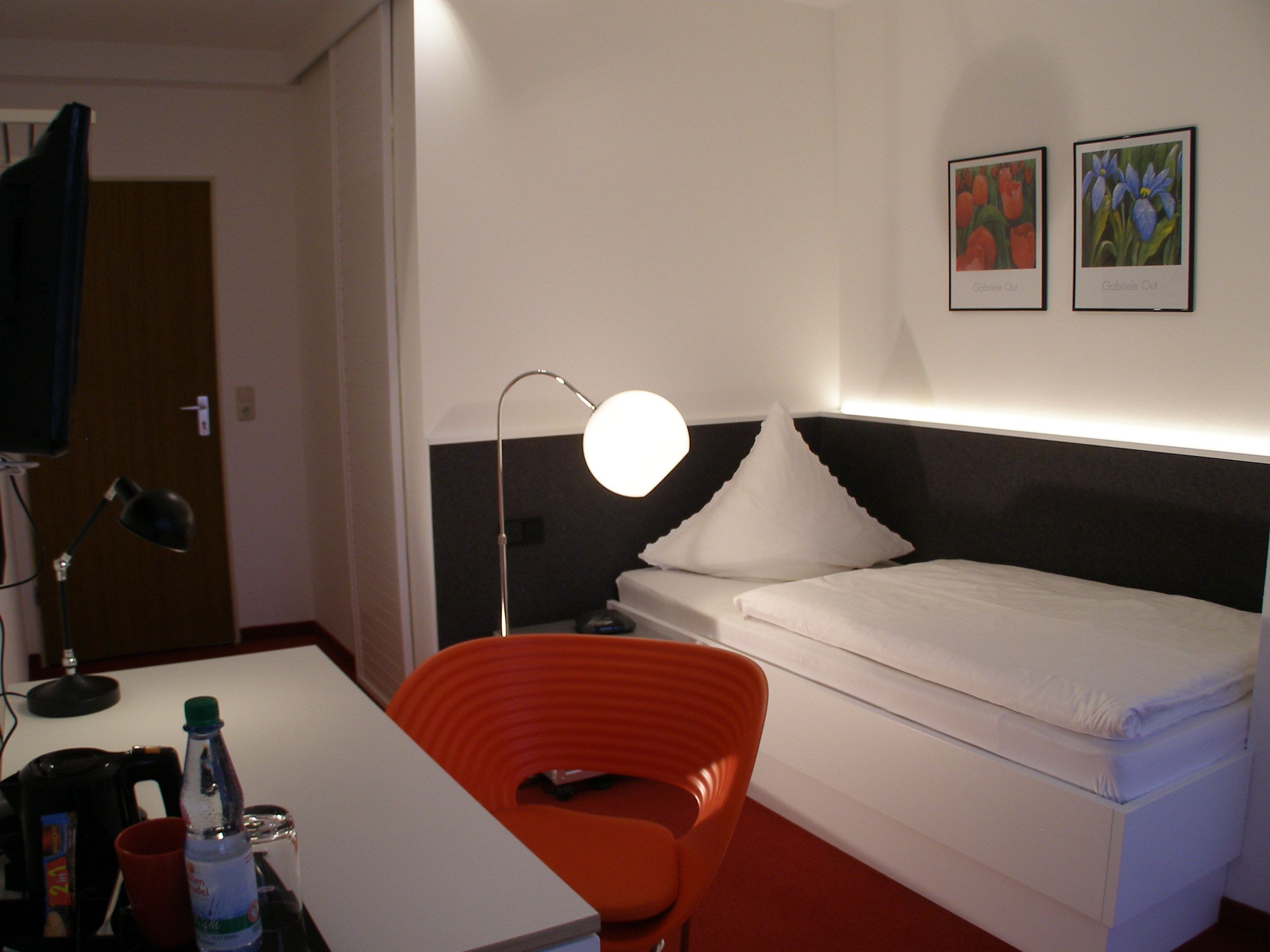 A hotel room with a bed, a desk with a lamp, a chair, and a nightstand with a bottle and glasses.