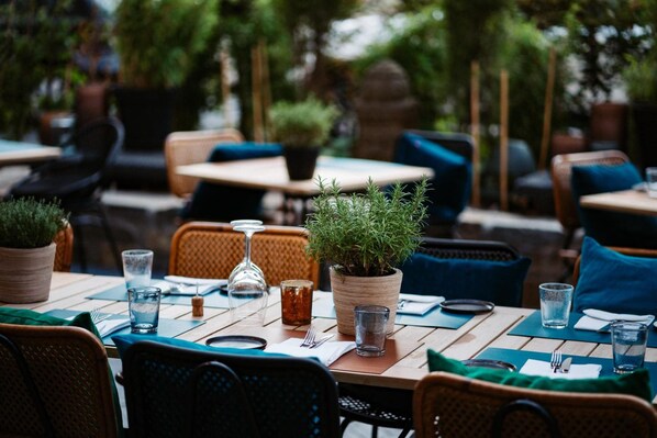Un área de comedor al aire libre con mesas preparadas para dos, plantas en macetas y cristalería.