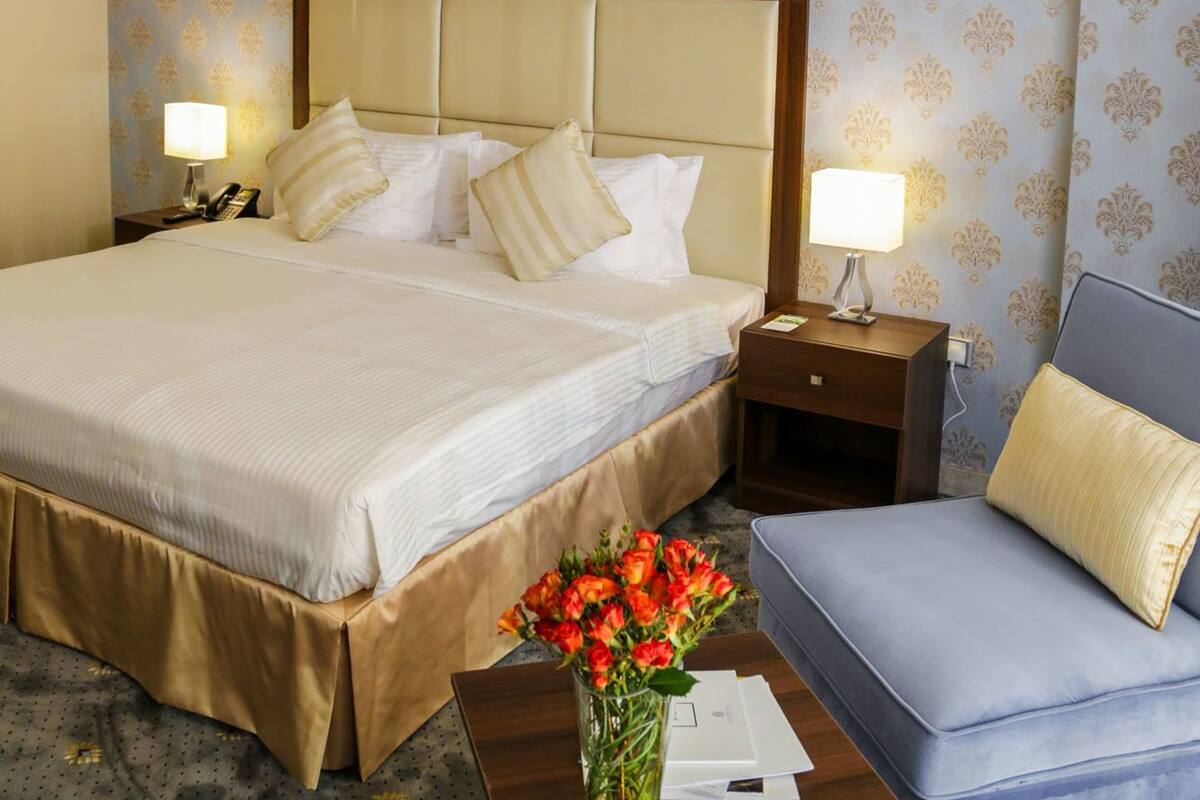 A hotel room with a large bed, a bedside table with a lamp, a small table with a vase of flowers, and a sofa.