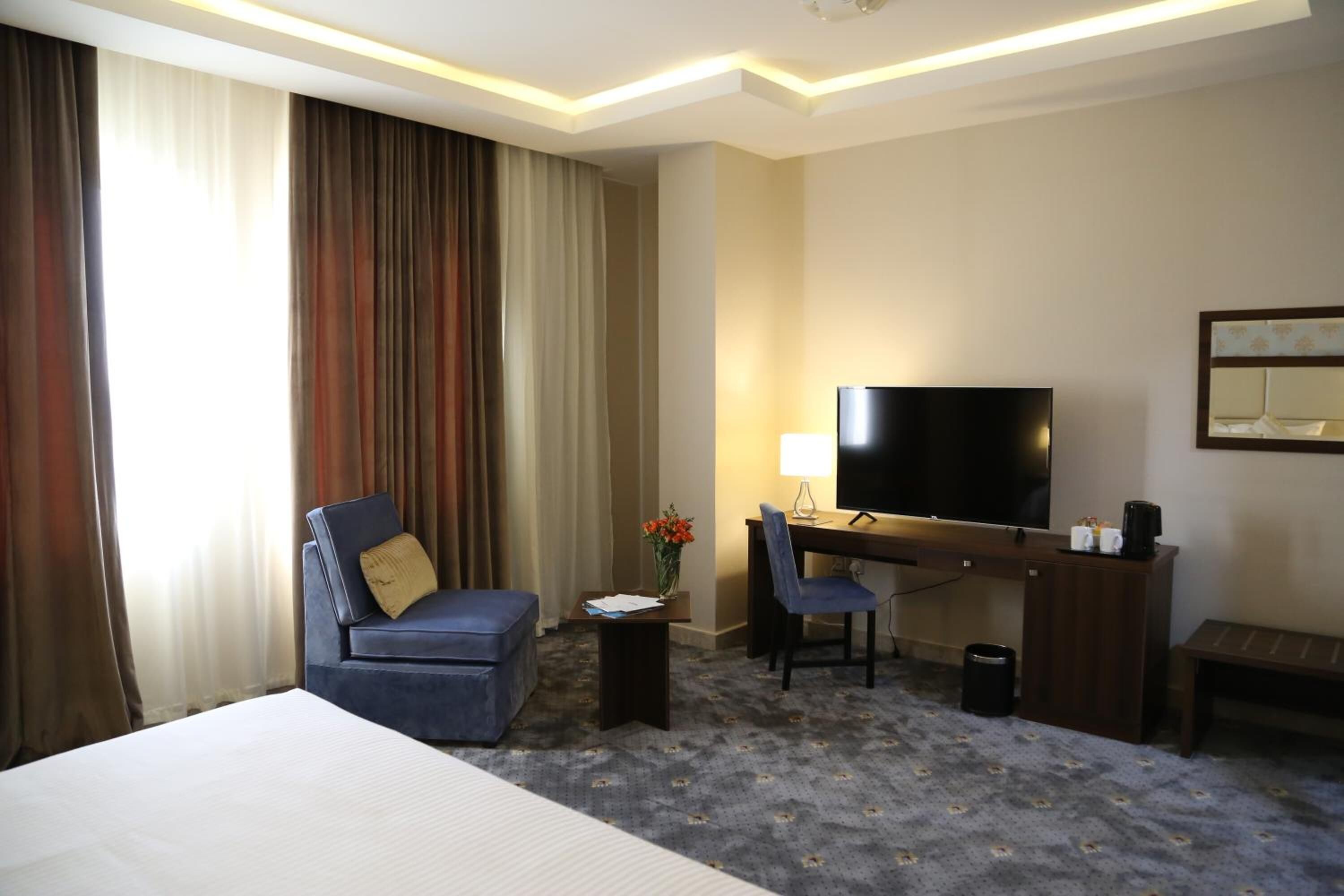 A hotel room with a bed, a desk with a TV, a chair, a sofa, a small table with a vase of flowers, and a carpeted floor.