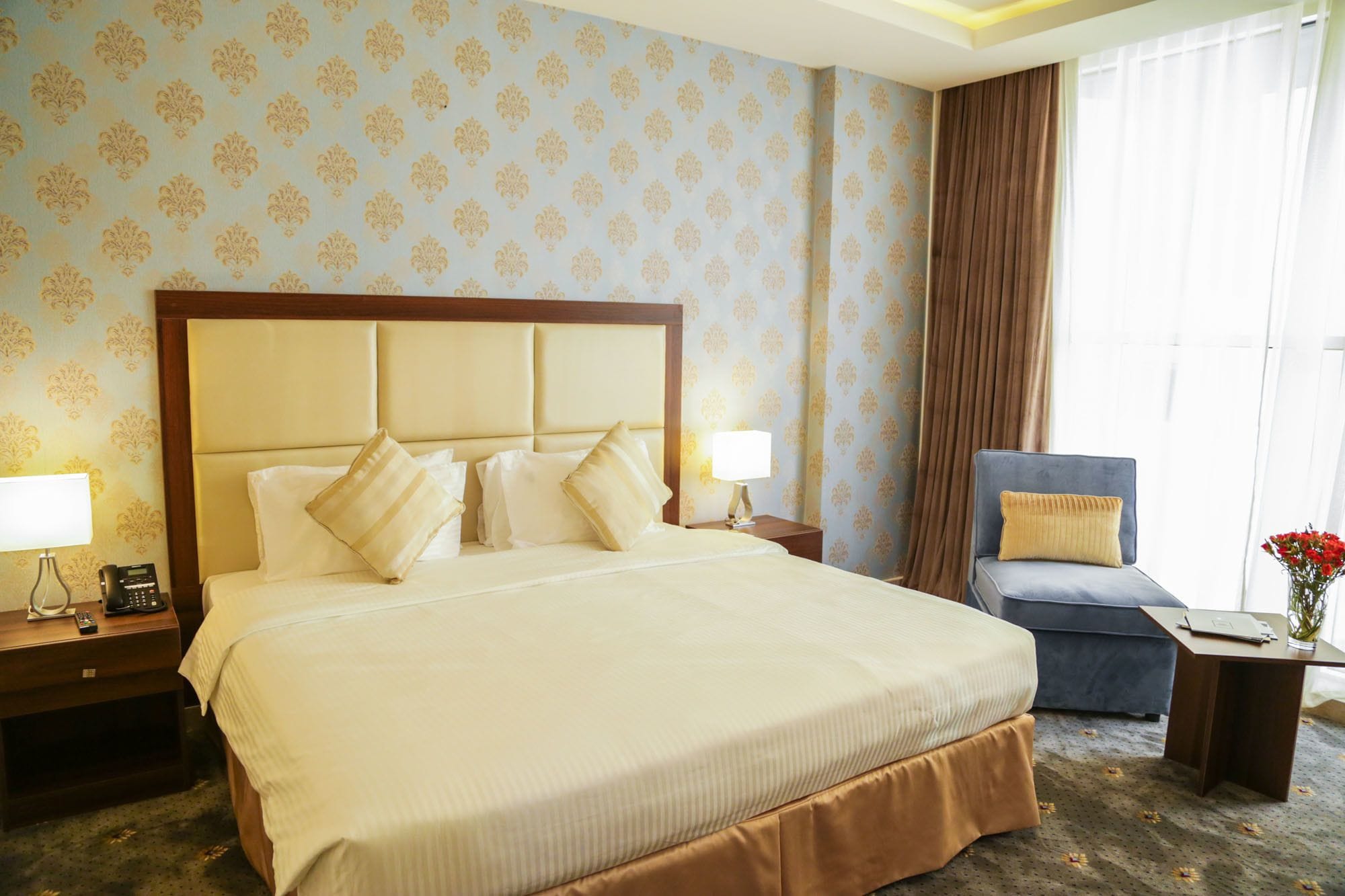 A hotel room with a large bed, a chair, a desk, and a vase with flowers.