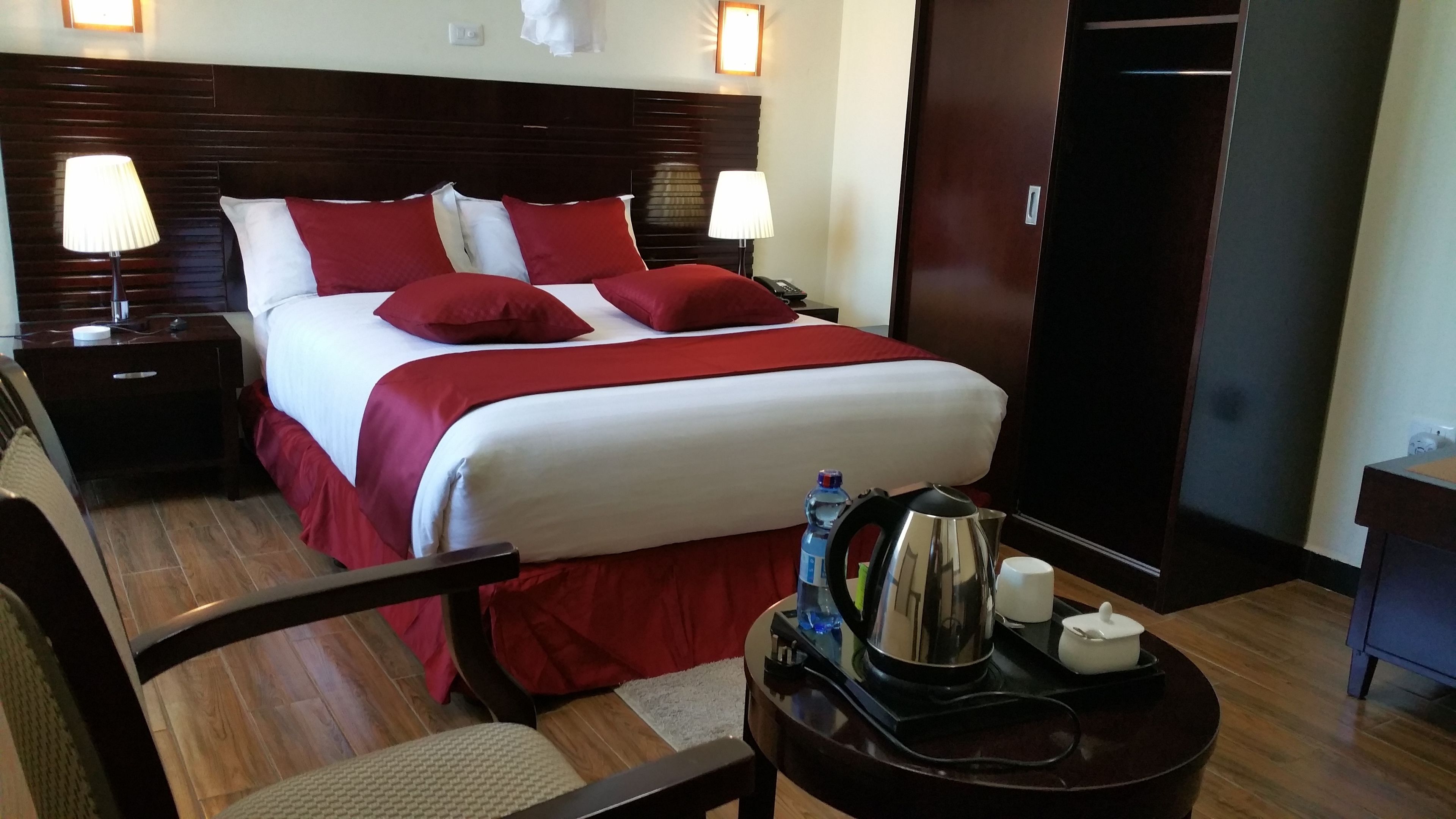 A hotel room with a large bed, two bedside lamps, a nightstand, a chair, a small table with a kettle and a bottle of water, and a door.