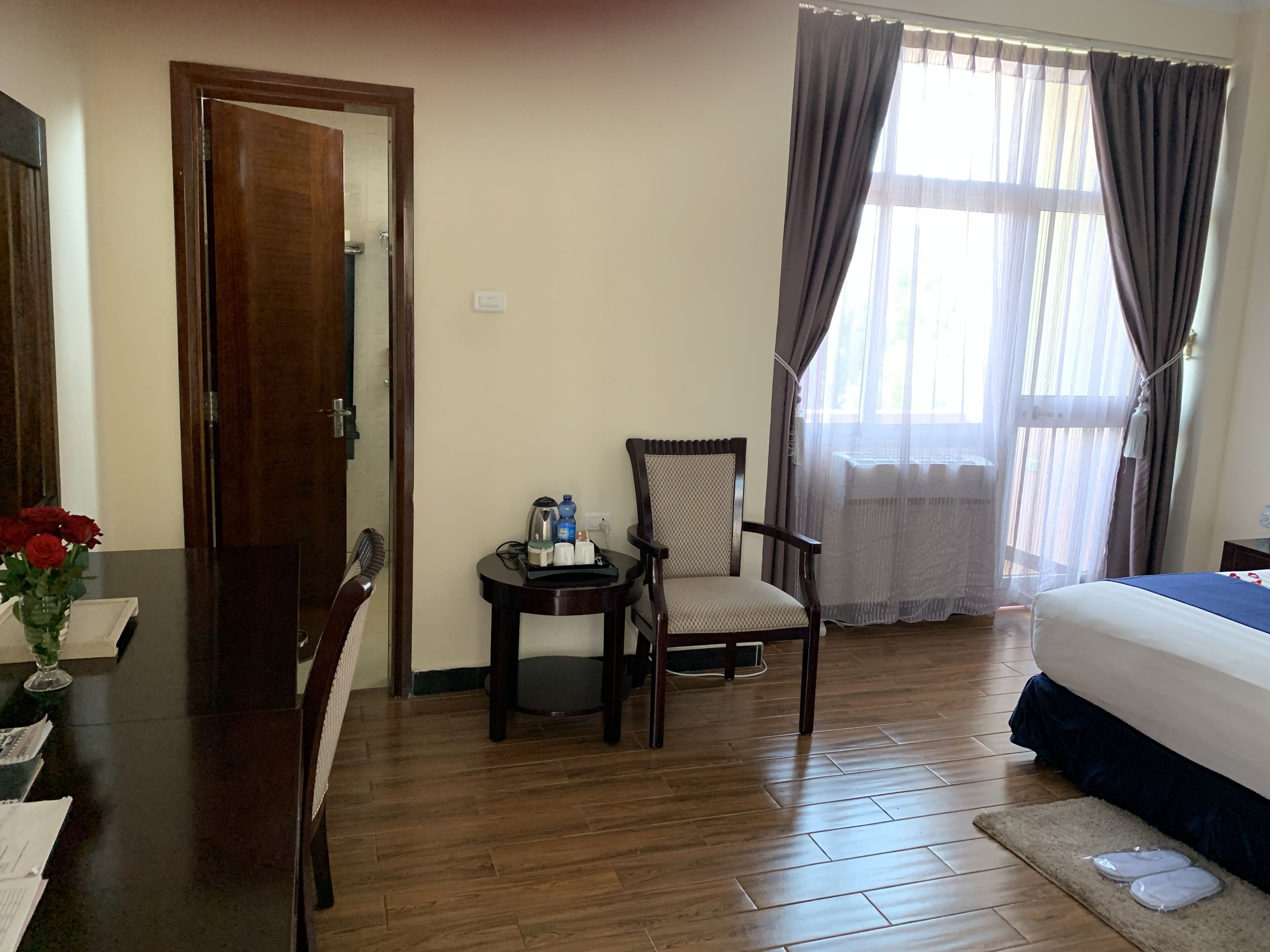A hotel room with a wooden floor, a bed, a chair, a small table with toiletries, and a window with curtains.