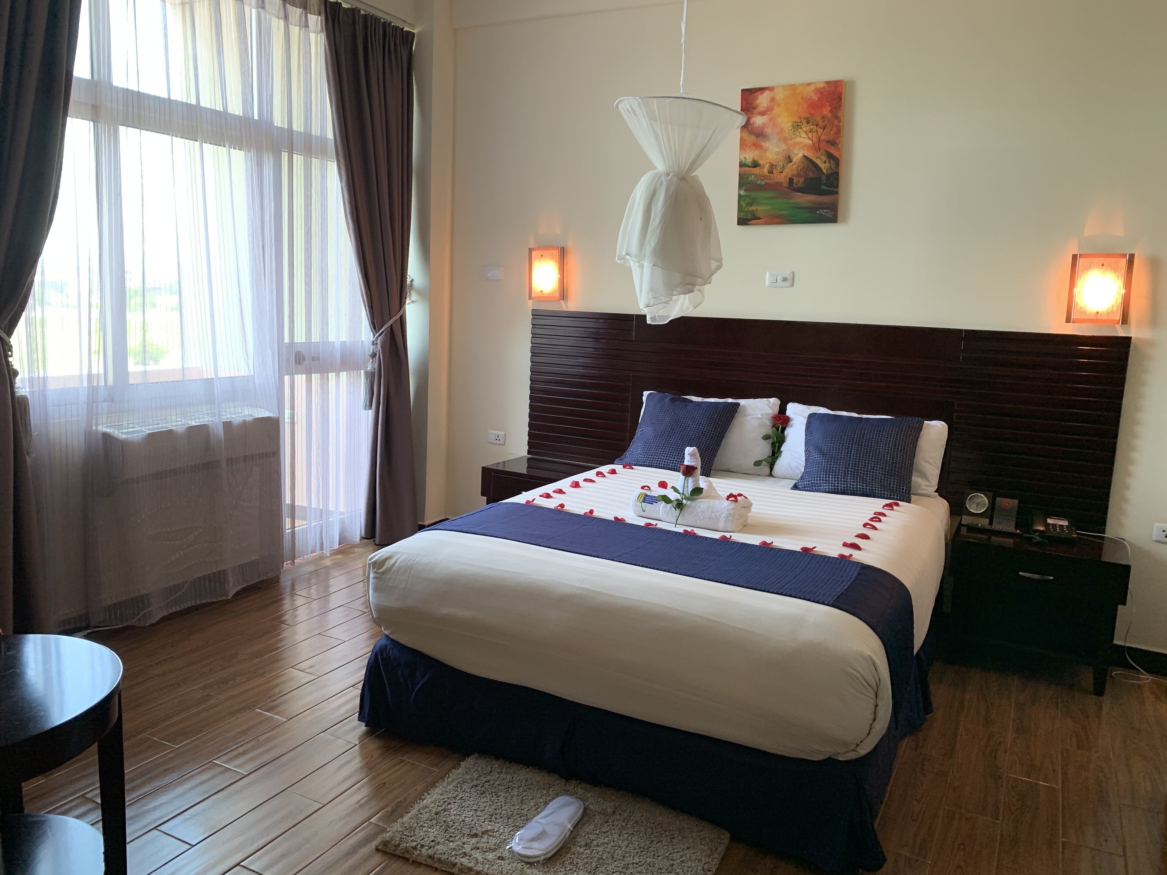 A neatly made bed with a blue and white bedspread, a wooden headboard, and a painting on the wall. There is a bedside table with a lamp and a clock.