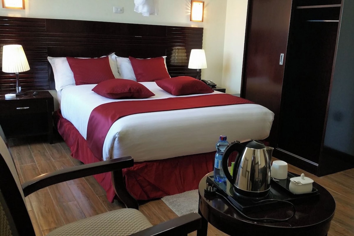A hotel room with a large bed, two bedside tables, a chair, a small table with a kettle and cups, and a door.
