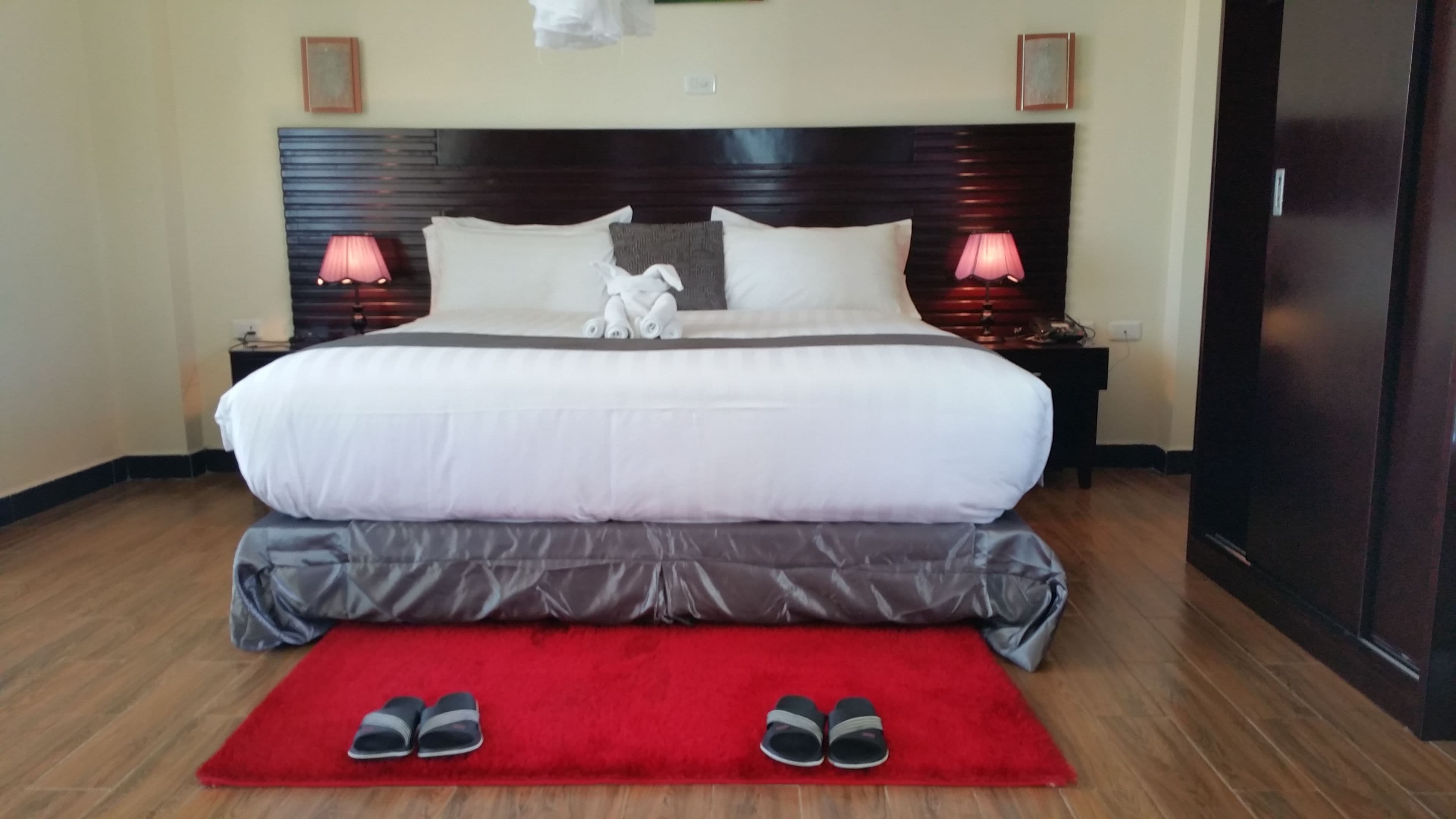 A neatly made bed with a red rug and slippers in front, wooden headboard, two bedside lamps, and a framed picture on the wall.