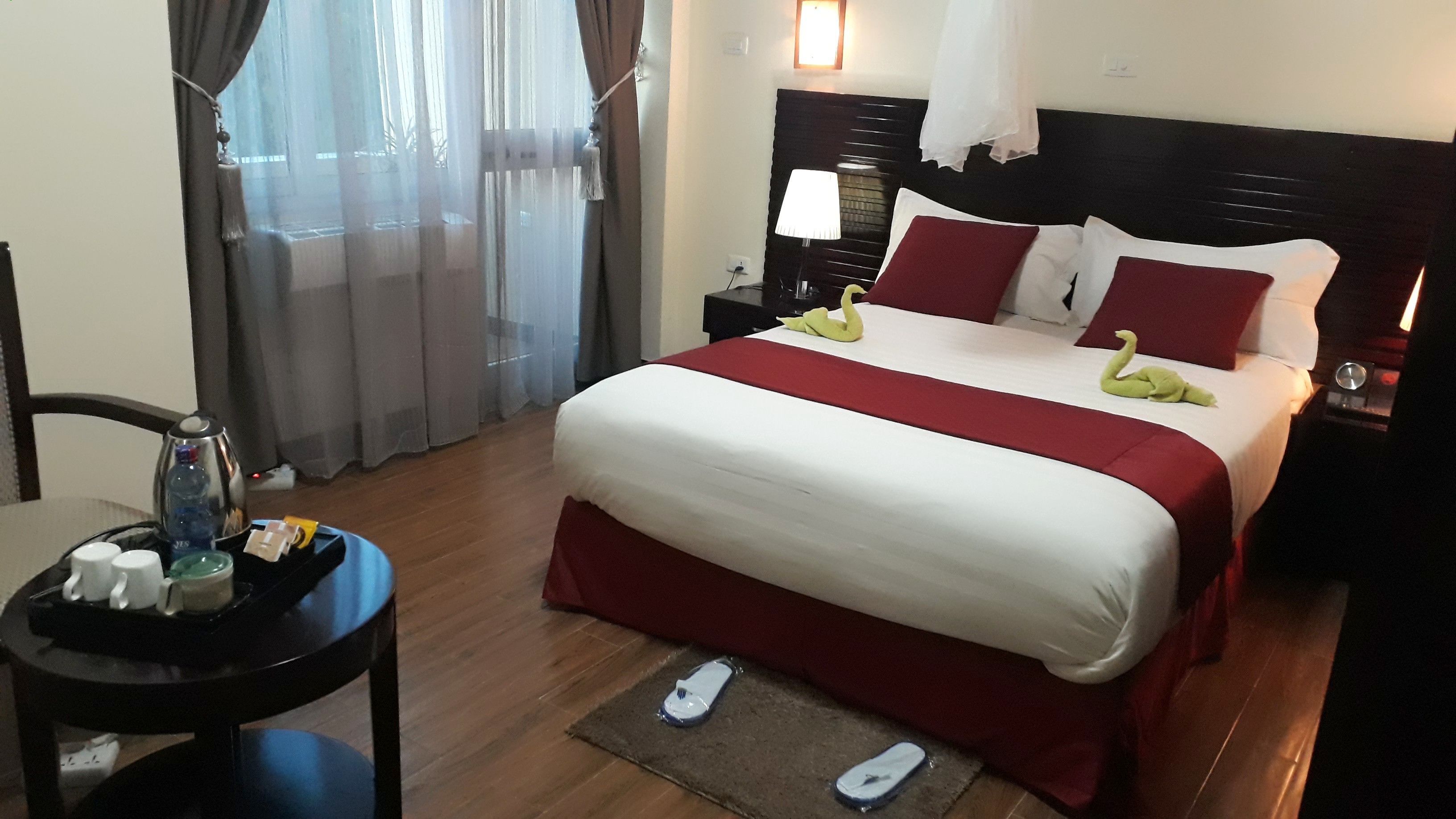 A hotel room with a large bed, a bedside table with a lamp, a nightstand, a chair, a small table with toiletries, and a window with curtains.