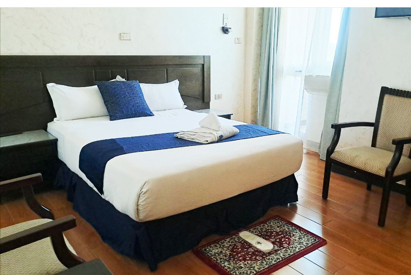 Une chambre d’hôtel avec un grand lit, une table de chevet, une chaise et un petit tapis.