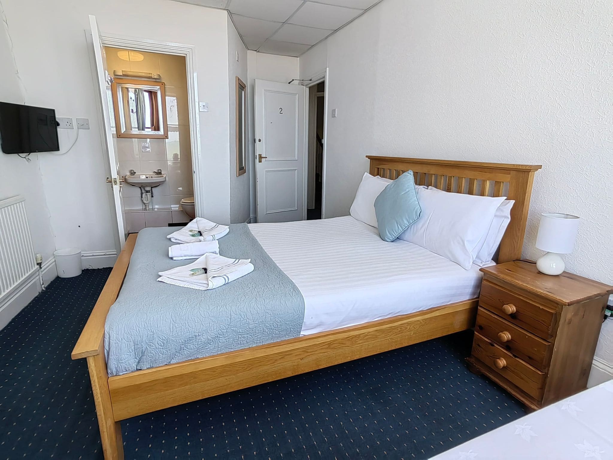A hotel room with a wooden bed, a bedside table with a lamp, a small dresser, a bathroom with a sink and mirror, and a TV mounted on the wall.