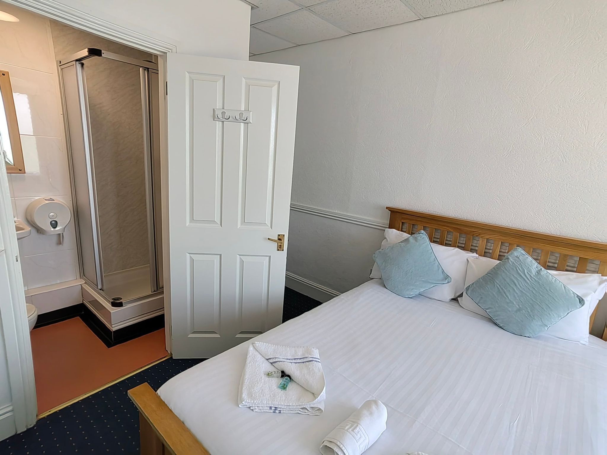 A hotel room with a bed, pillows, a towel, a wooden headboard, a white door, and a shower area.