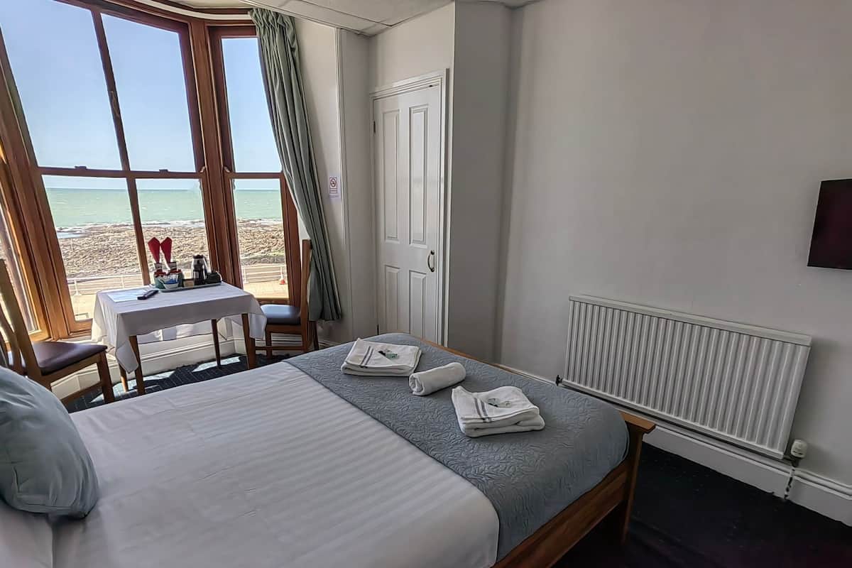 A hotel room with a bed, a table with a chair, a view of the beach, and a white door.