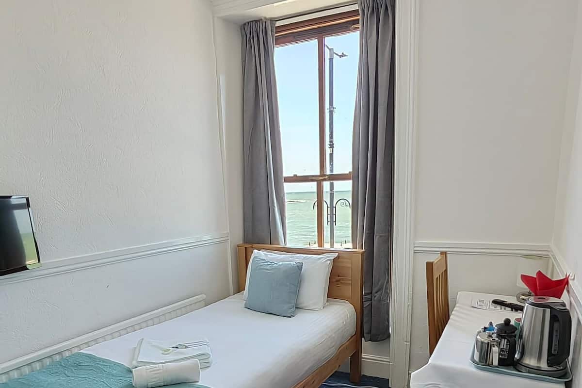 A small hotel room with a single bed, a desk with a chair, a window with curtains, and a view of the sea.