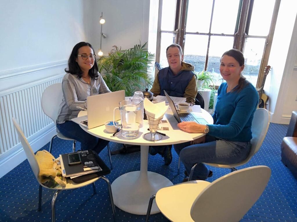 Eine Gruppe von Menschen sitzt an einem runden Tisch mit Laptops, Papieren und einem Glas Wasser. Es gibt Stühle mit gelben Kissen, eine Topfpflanze und eine Gitarre im Hintergrund.