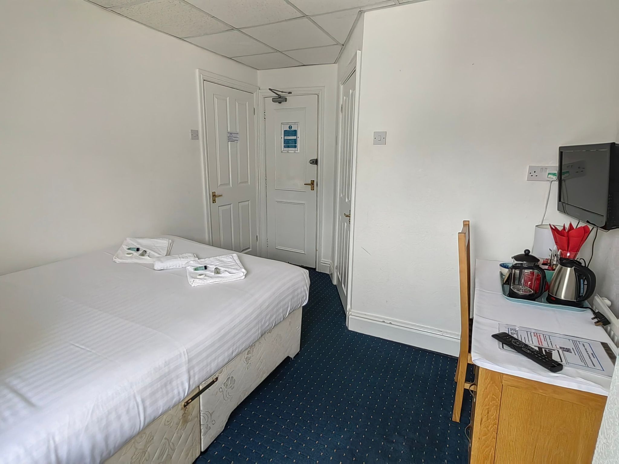 Une chambre d’hôtel avec un lit simple, un petit bureau avec une bouilloire, une télévision et une porte donnant sur une autre chambre.