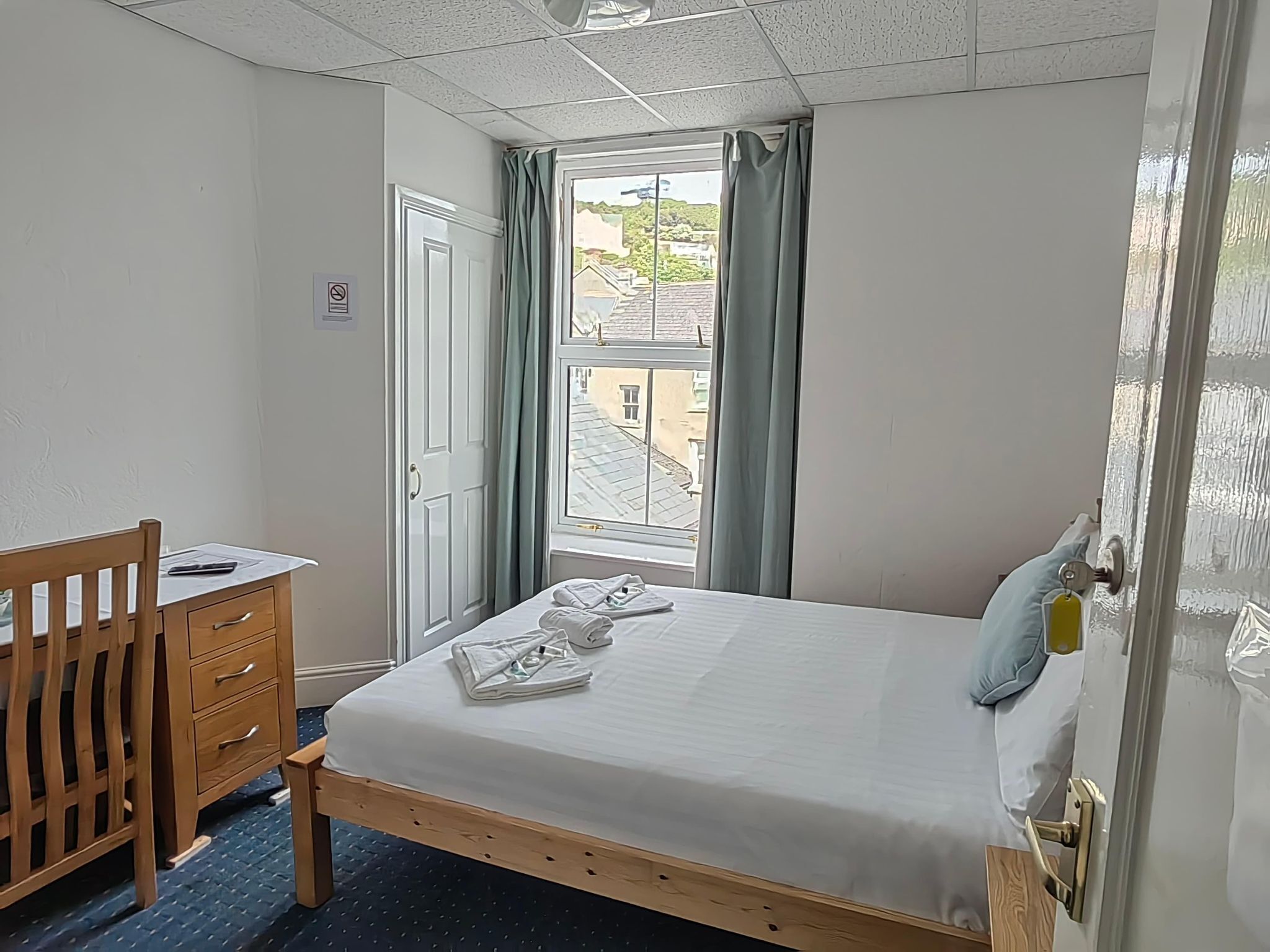 A bedroom with a bed, a wooden desk, a chair, a window with curtains, and a view of buildings outside.