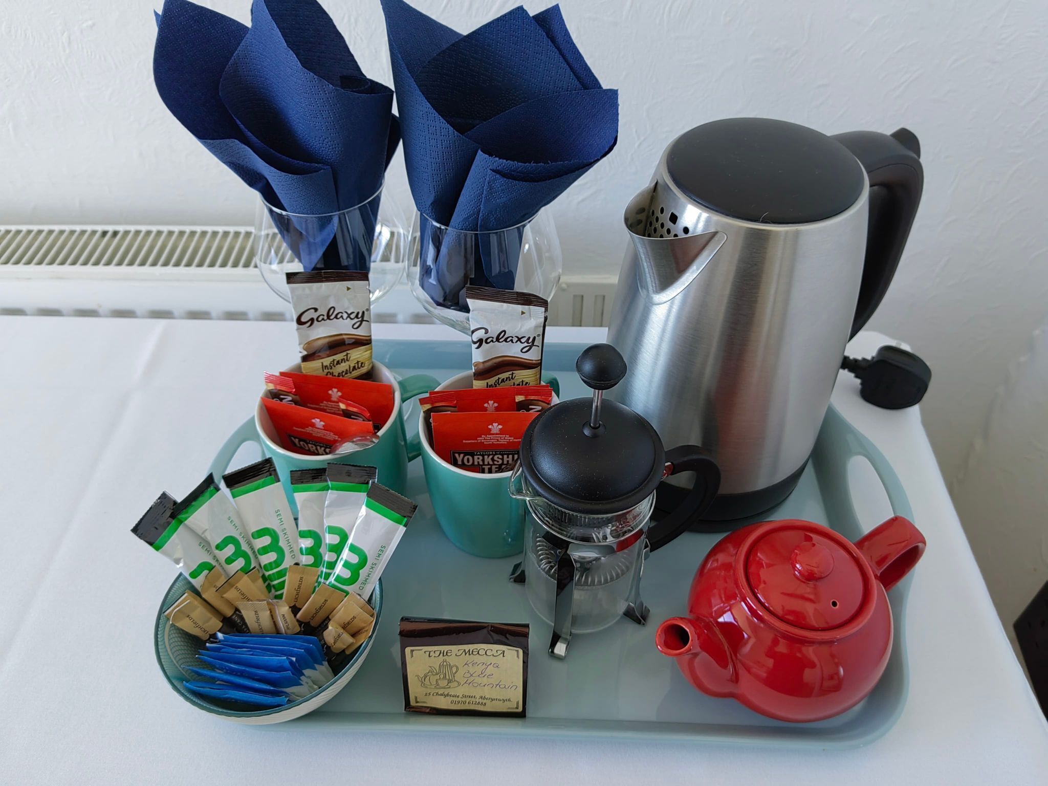 Une table avec une machine à café, une théière rouge, une carafe en verre et divers en-cas emballés.