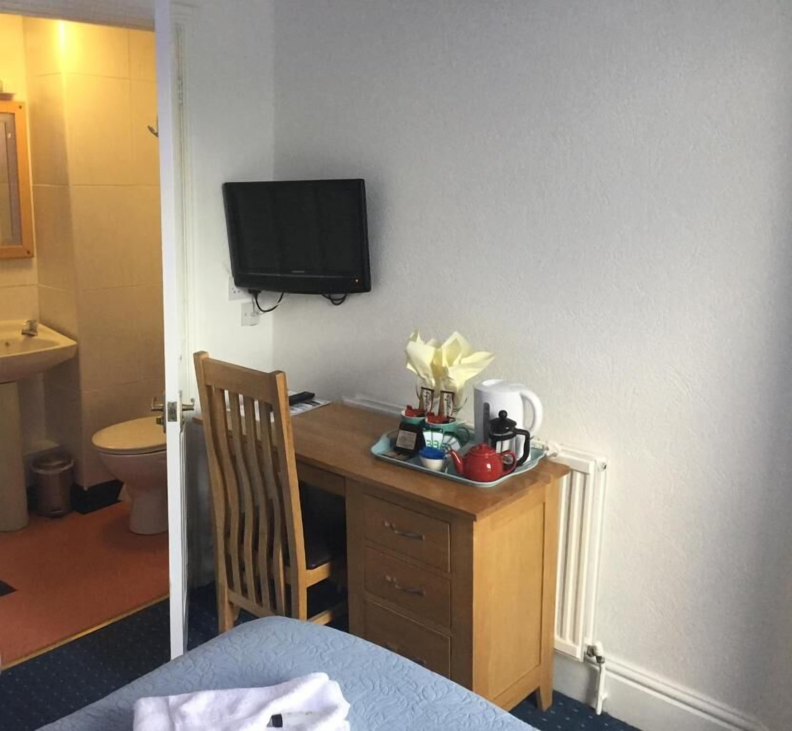 A hotel room with a bed, a wooden desk with a kettle, a television, and a small bathroom with a toilet and sink.