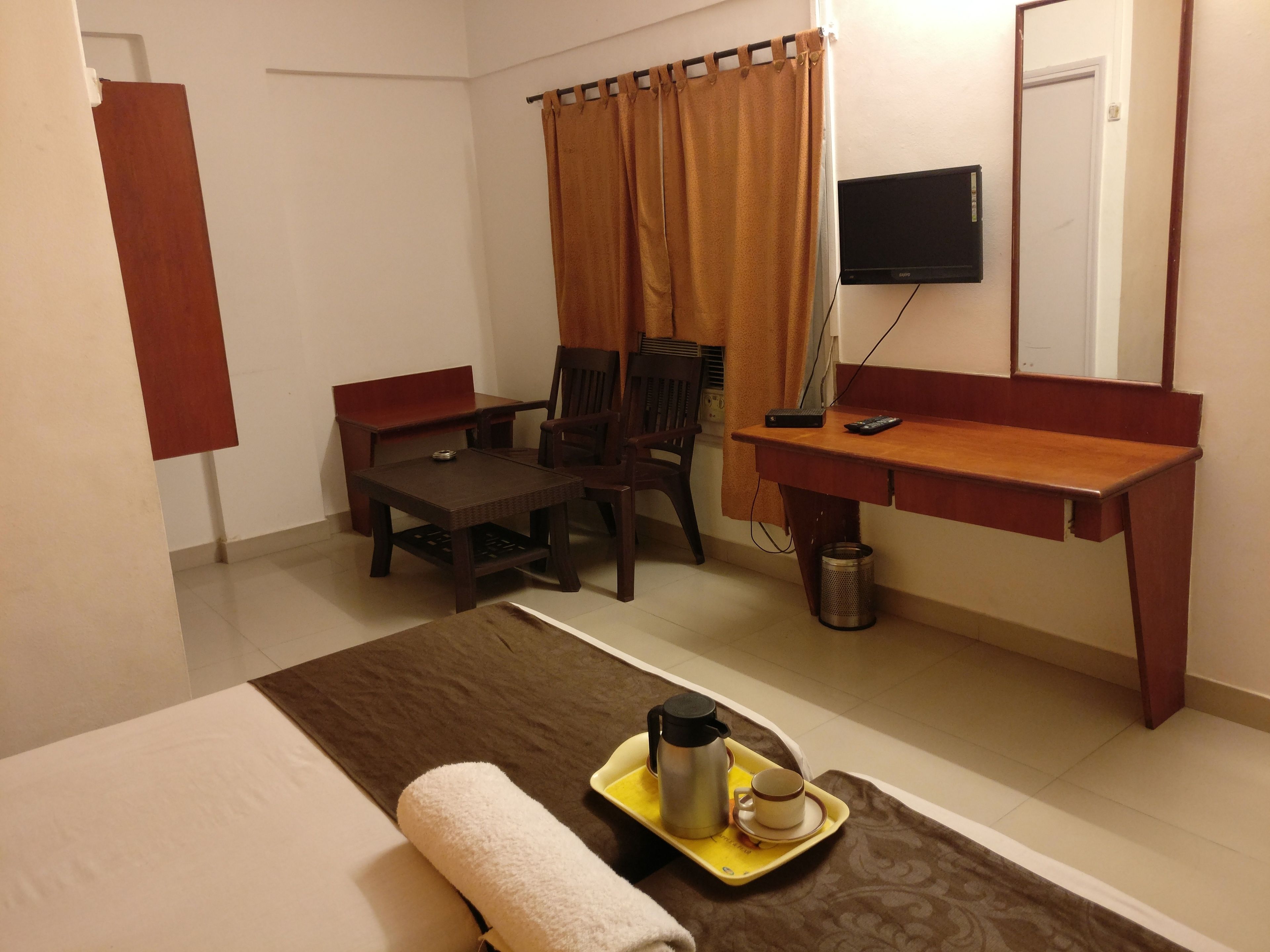 A hotel room with a bed, a desk, a chair, a TV, and a tray with a cup and a thermos.