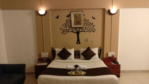 A neatly made bed with a tree decal on the wall, a bedside table with a phone and a lamp, and a bench in front of the bed. - HMR Royal Inn (Pune)