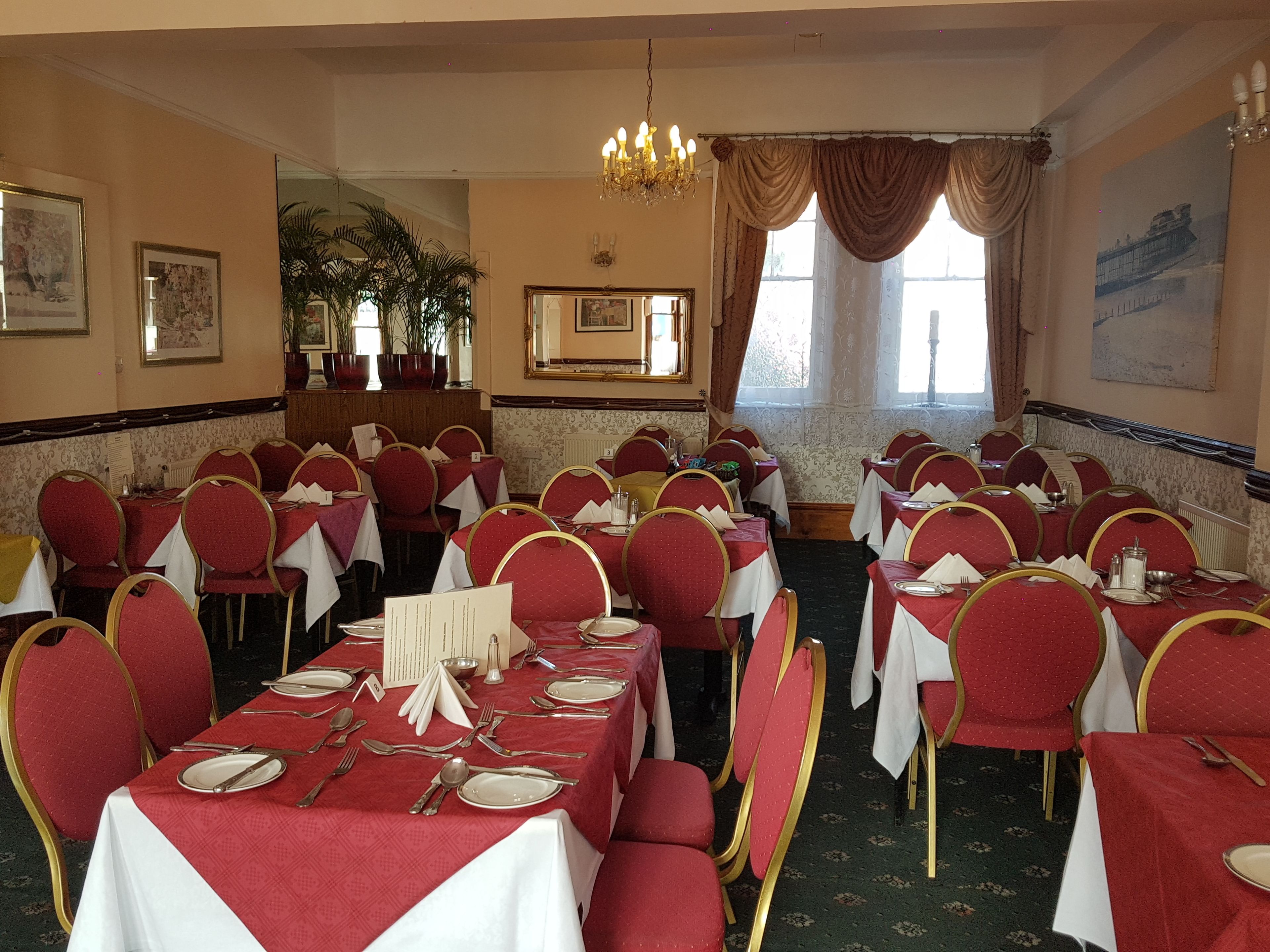 Una sala de banquetes amb taules rodones preparades per dinar, cadires tapissades de color vermell i un penjador.