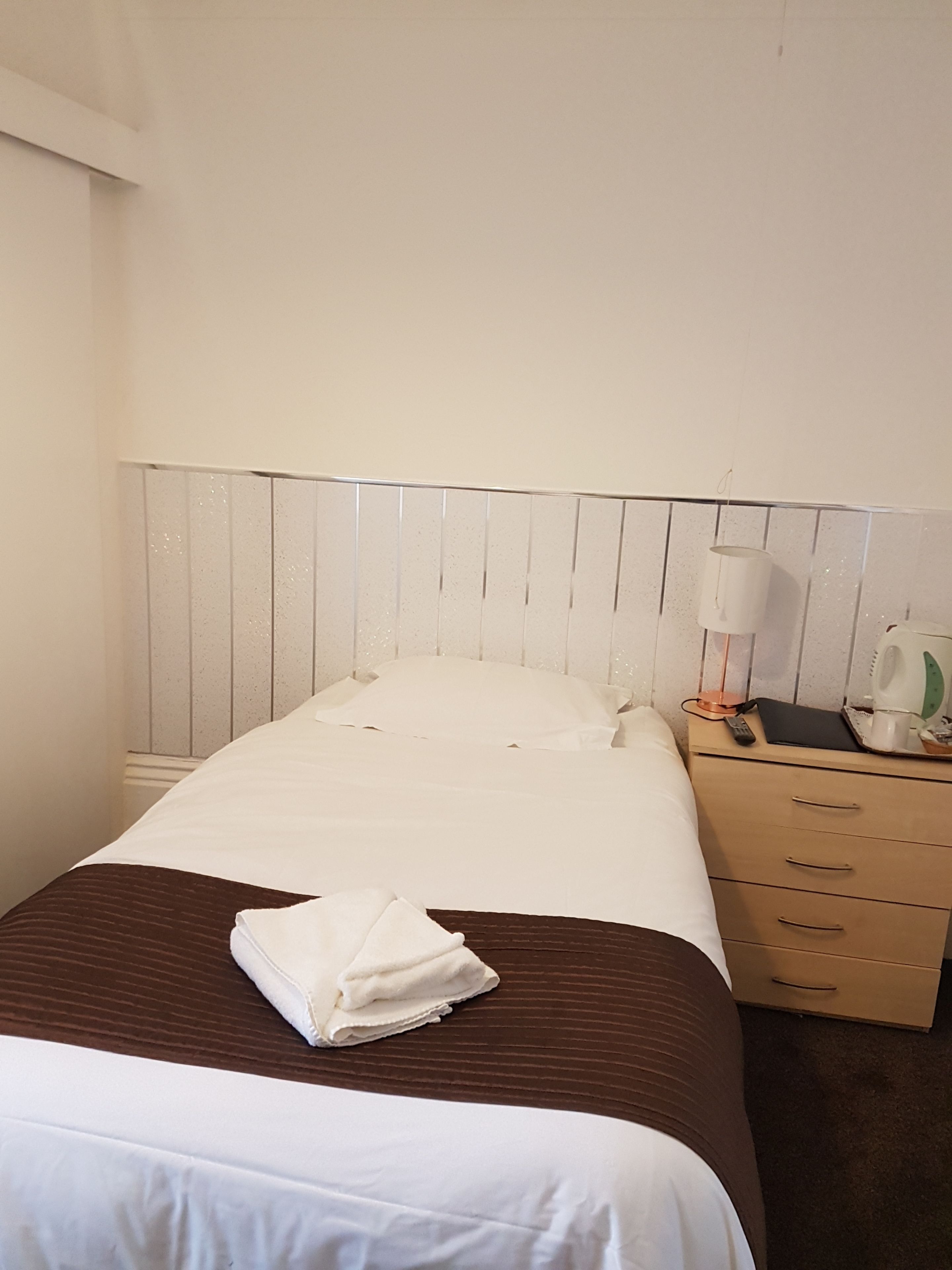 Una cama bien hecha con sábanas blancas, una toalla doblada y una mesita de noche con una lámpara y un control remoto.