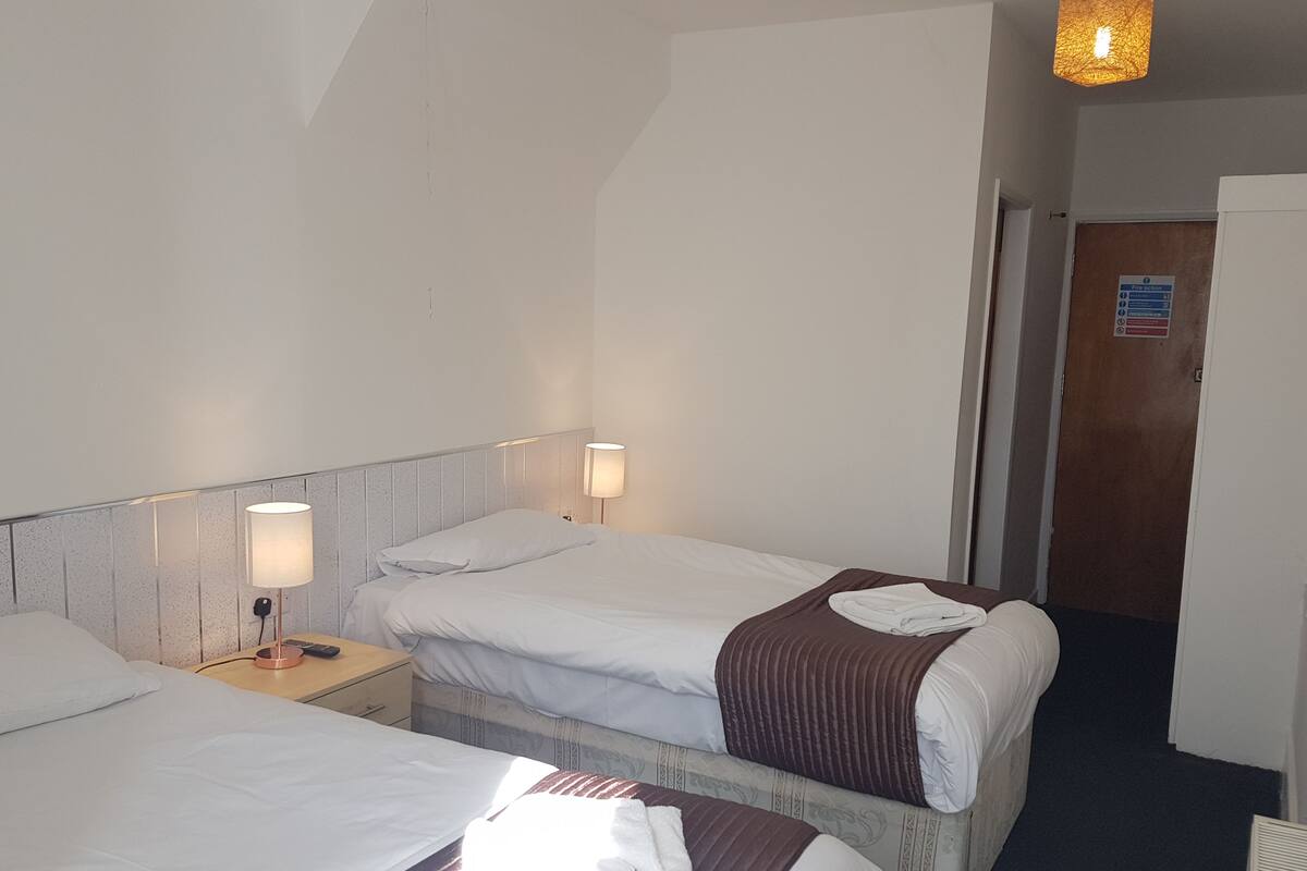 A hotel room with two beds, each with white linens and a brown blanket. There is a nightstand with a lamp and a power outlet. A door with a sign is visible in the background.