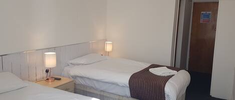 A hotel room with two beds, each with white linens and a brown blanket. There is a nightstand with a lamp and a power outlet. A door with a sign is visible in the background.