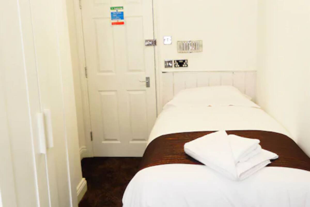 A small, single-bed room with a white door, a small window, and a radiator on the wall.