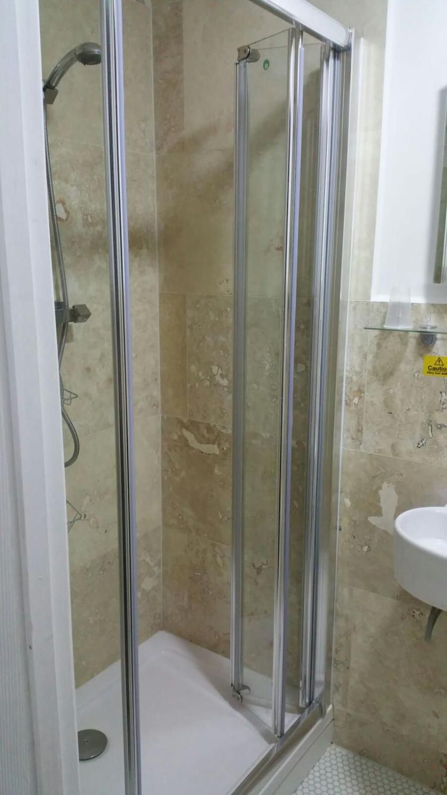 A glass-enclosed shower with a handheld showerhead, a white sink, and a caution sign on the wall.