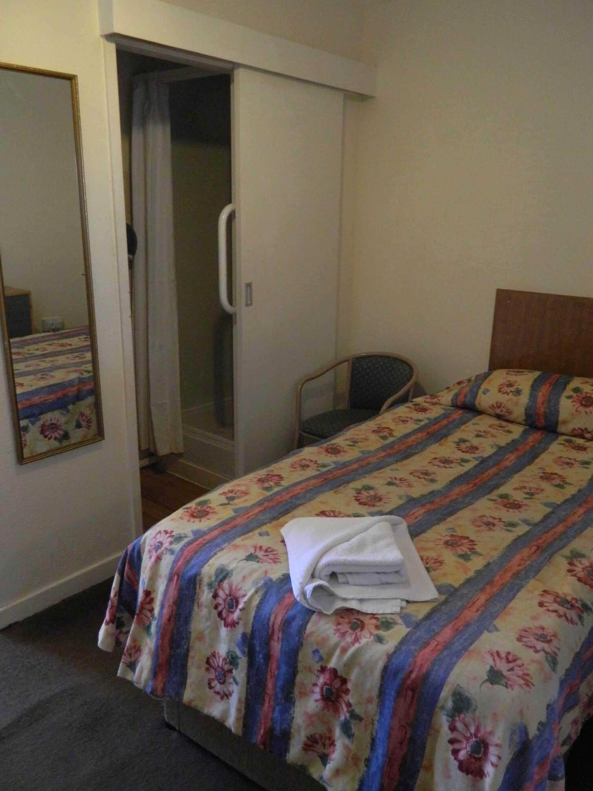 Una habitación de hotel con una cama, una silla, zona de ducha y un espejo.